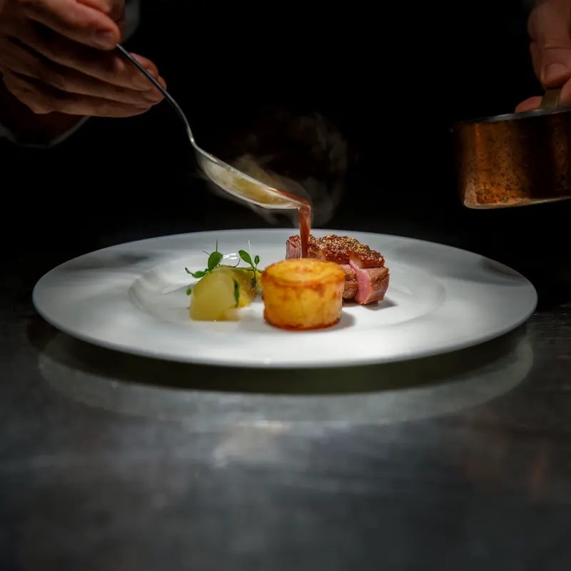 Koch gießt dampfende Sauce über ein angerichtetes Gericht